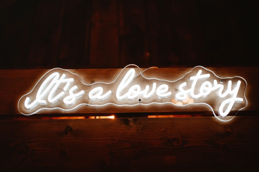 A white neon sign shines brightly on a dark wooden wall, with the text "Its a love story".