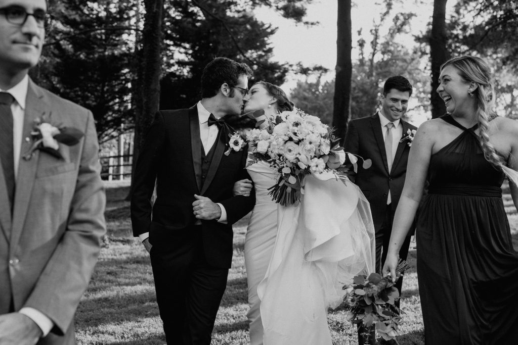 Bride and Groom kiss while walking, surrounded by their wedding party. Black and white image.  Outdoors on the grounds of The Barns at Wolf Trap.