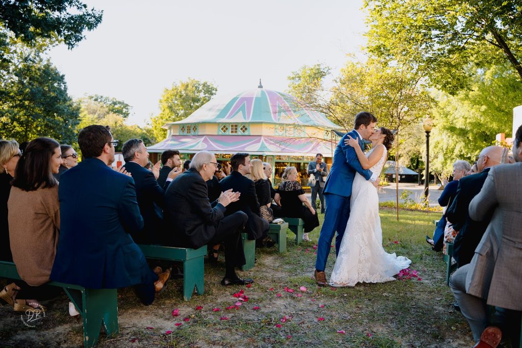 Bride and Groom embrace after their vows while their guests look on and applaud.  A modern chuppah and a historic carousel are in the background at Glen Echo.