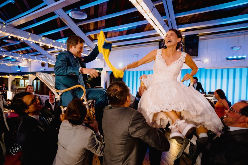 Bride and Groom laughing in lifted chairs for the Hora, supported by an energetic crowd, in the Bumper Car Pavilion at Glen Echo.