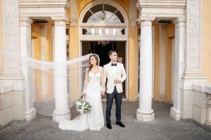 Bride and Groom stand outside of a formal building, looking away from each other. Bride's long veil is being blown horizontally to the edge of the frame. Photography @joyphoto